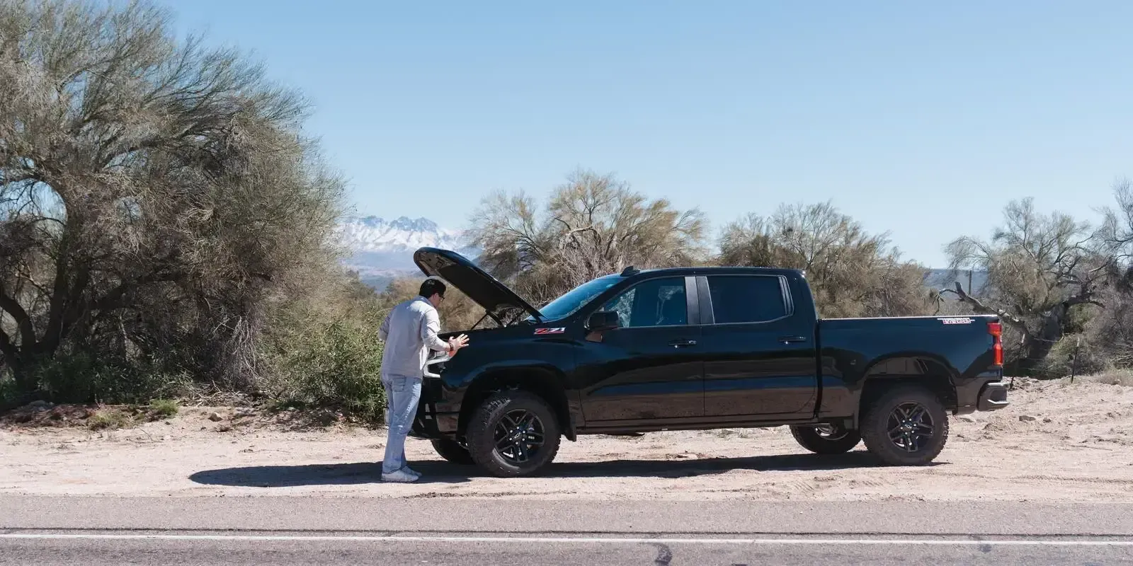 A man on the side of the road looking under the hood of his truck in need of roadside assistance.