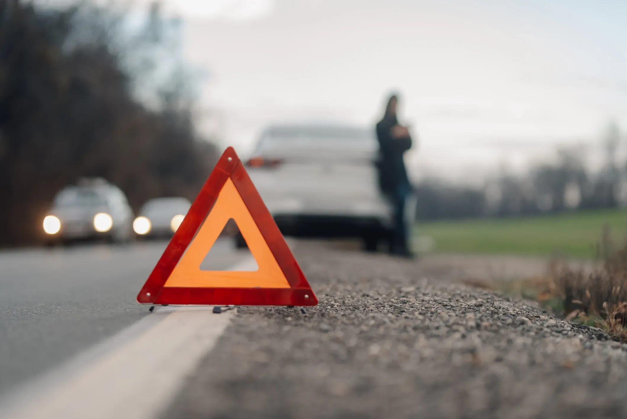 An orange safety triangle with a blurred person and car pulled over in the background.