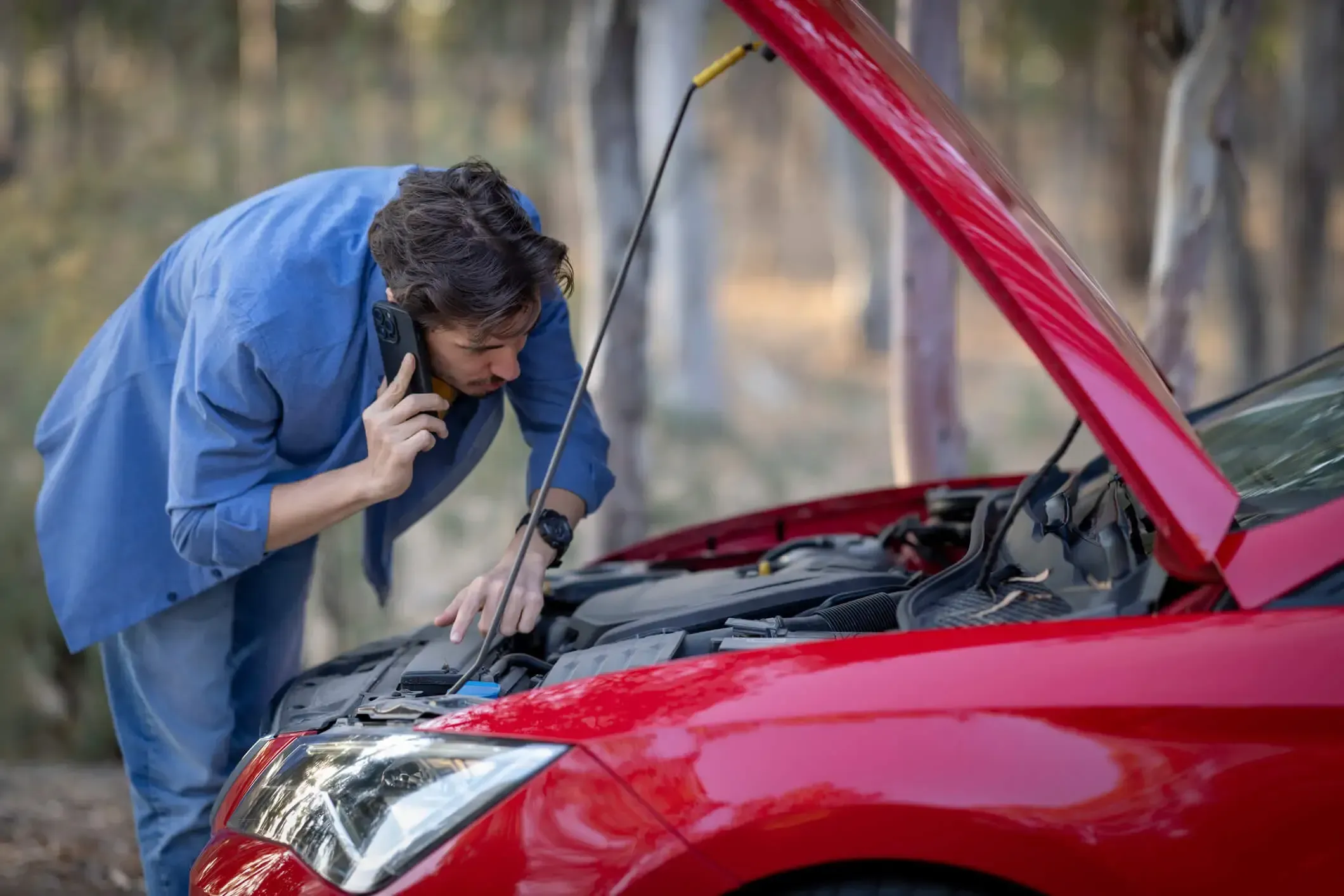 A man looking under the hood of his red car calling for roadside assistance.