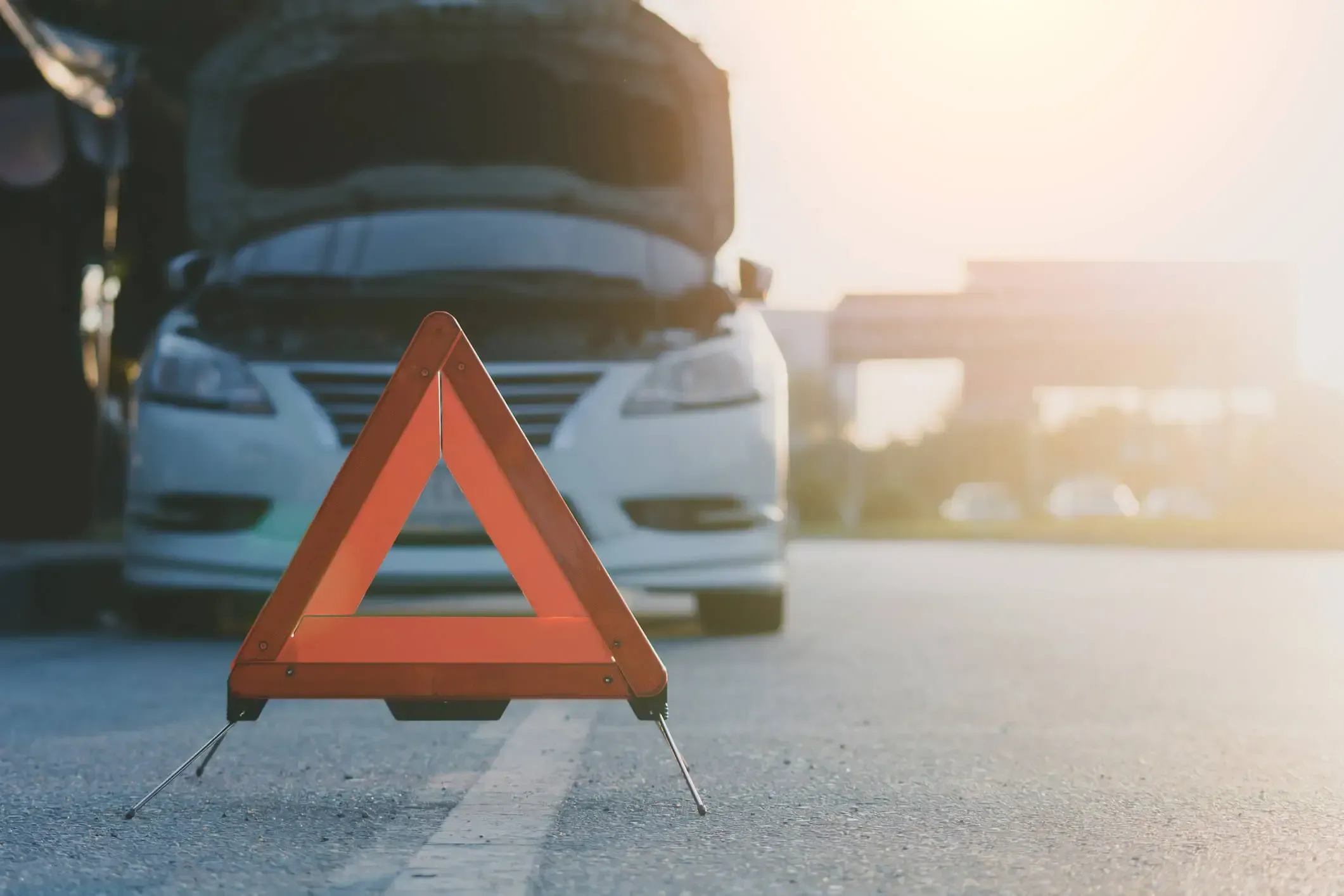 An orange emergency safety triangle on the side of the road with a car with its hood up in the background.
