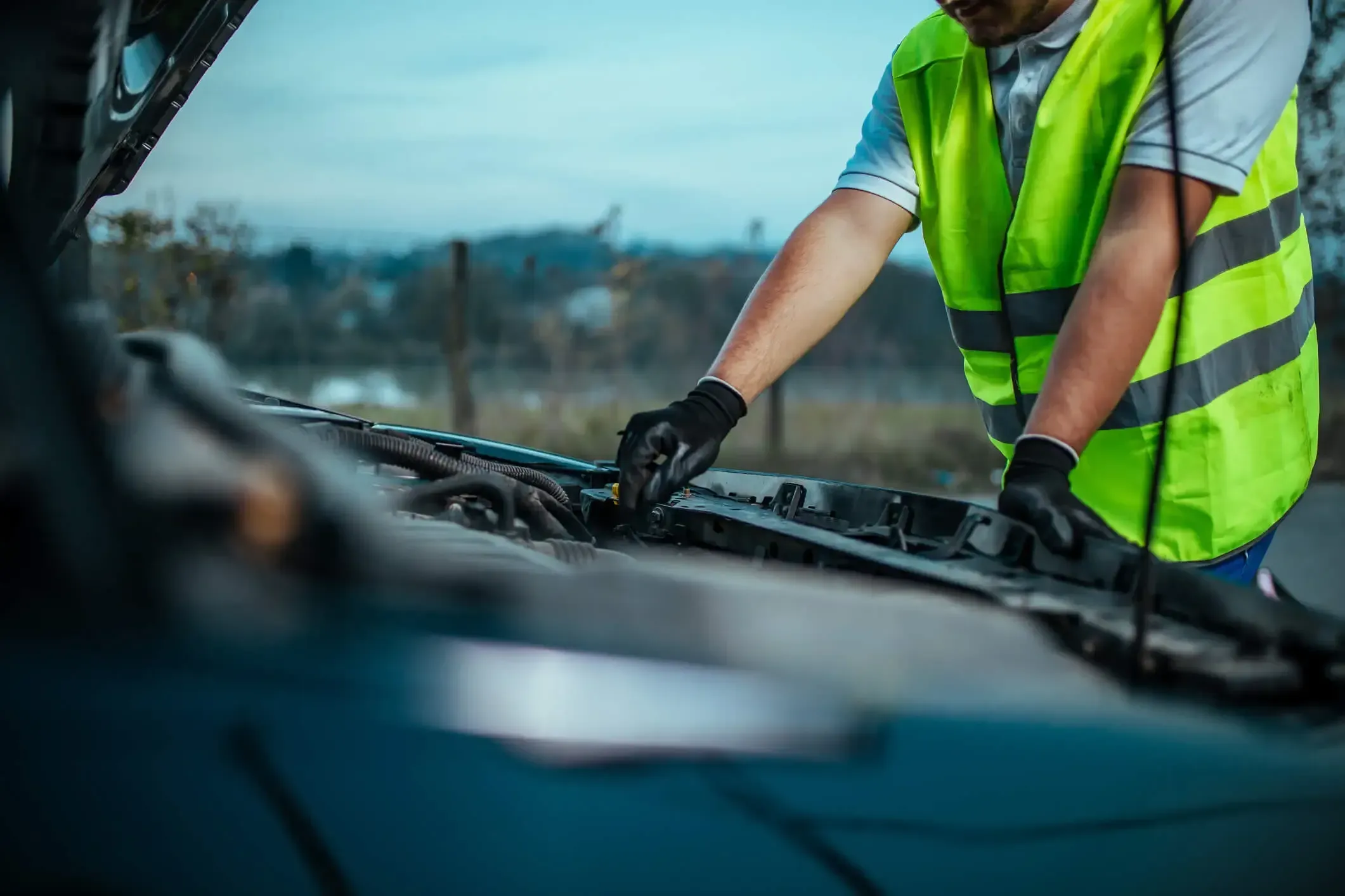 A roadside assistance worker in a high visibility safety vest looking under the hood of a car.