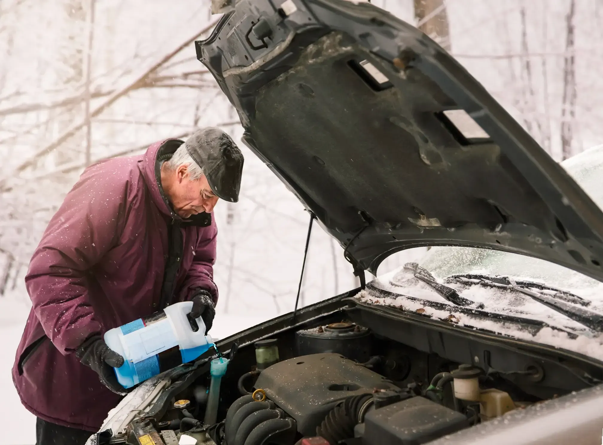 An elderly man outside in the winter with his car's hood up performing winter maintenance by refilling fluids.
