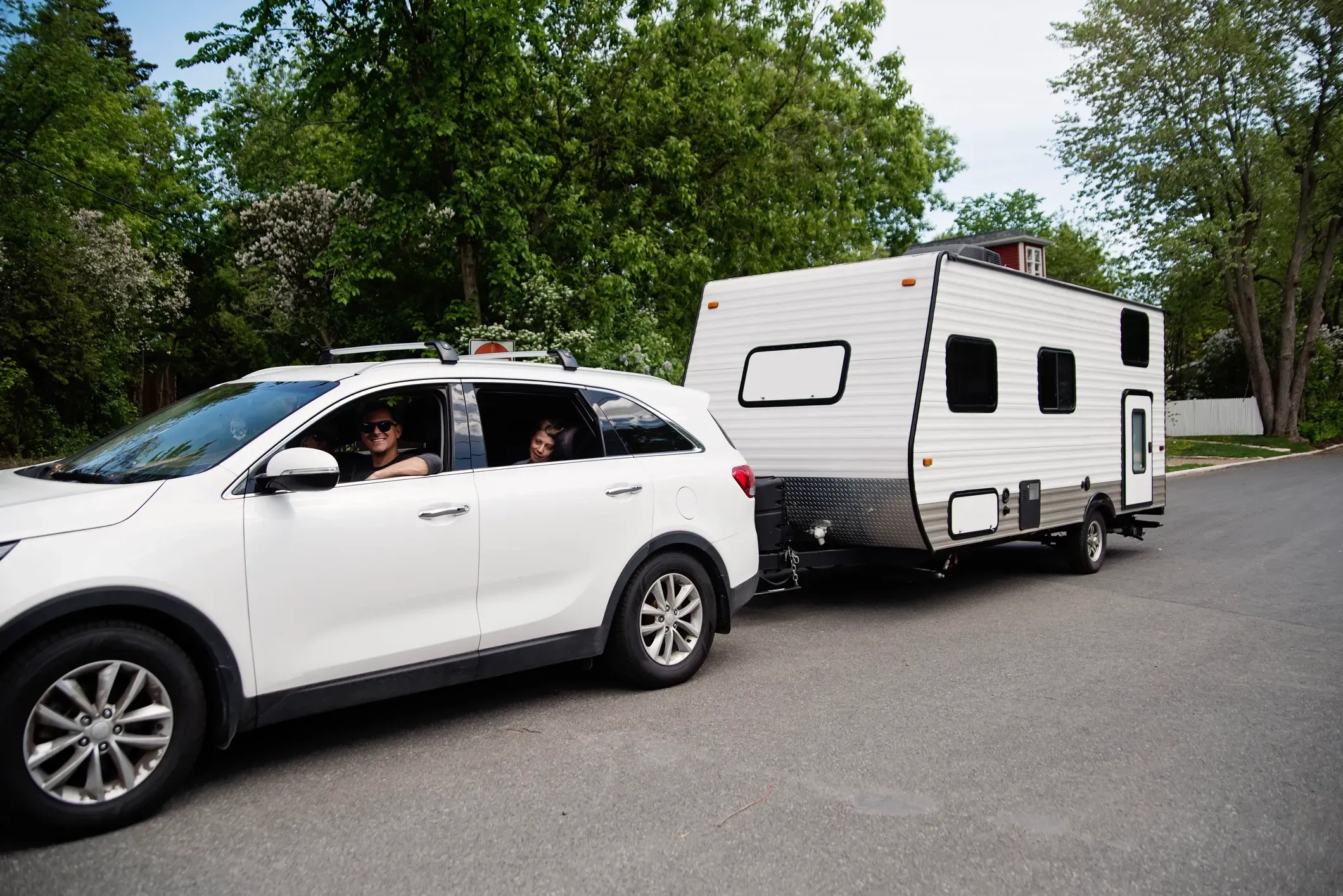 A family in a white compact SUV towing their travel trailer through their neighborhood on a camping trip.