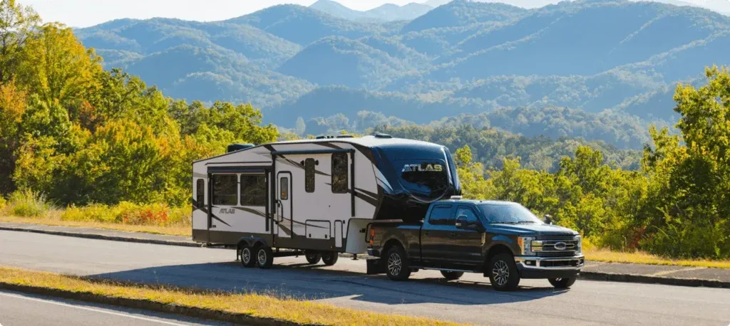 A truck tows an Atlas travel trailer with lush green rolling mountains in the background.