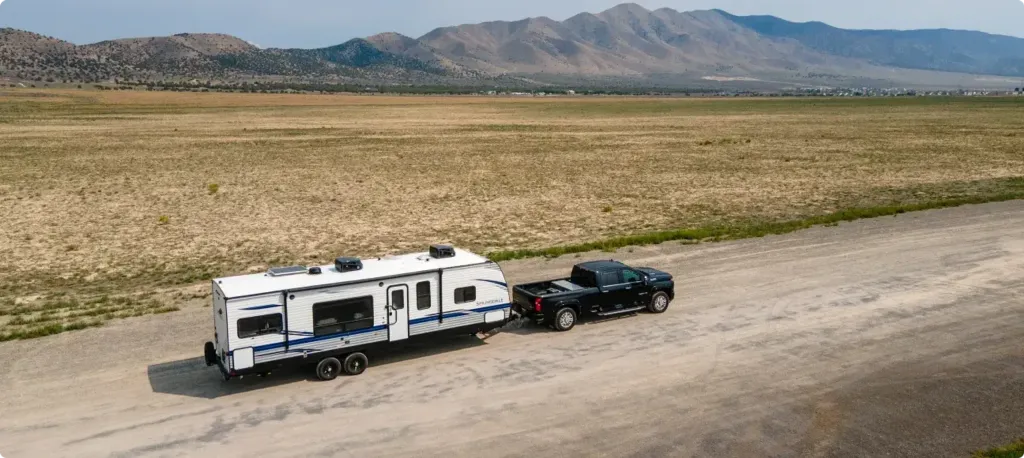 A truck tows a fifth wheel in front of a wide dusty field with tall rocky mountains in the background