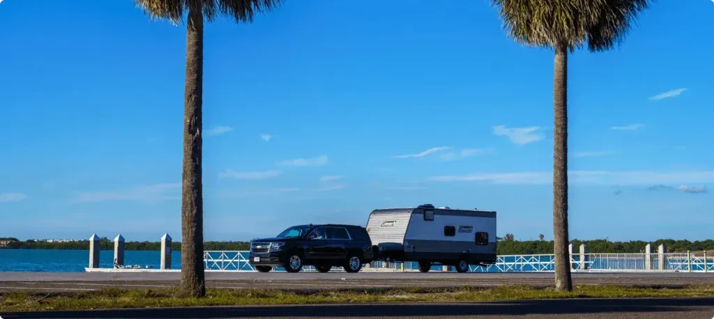 An SUV pulls a Coleman trailer along a road with a coastal waterway in the background.