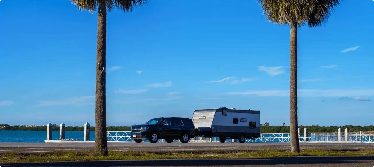 An SUV pulls a Coleman trailer along a road with a coastal waterway in the background.
