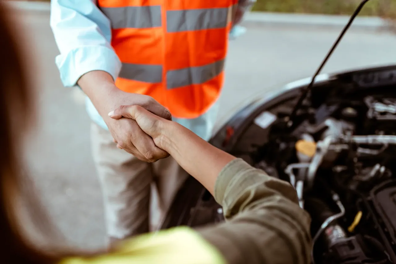 A roadside assistance technician wearing an orange safety vest shaking the hand of an individual over a vehicle with the hood popped up.
