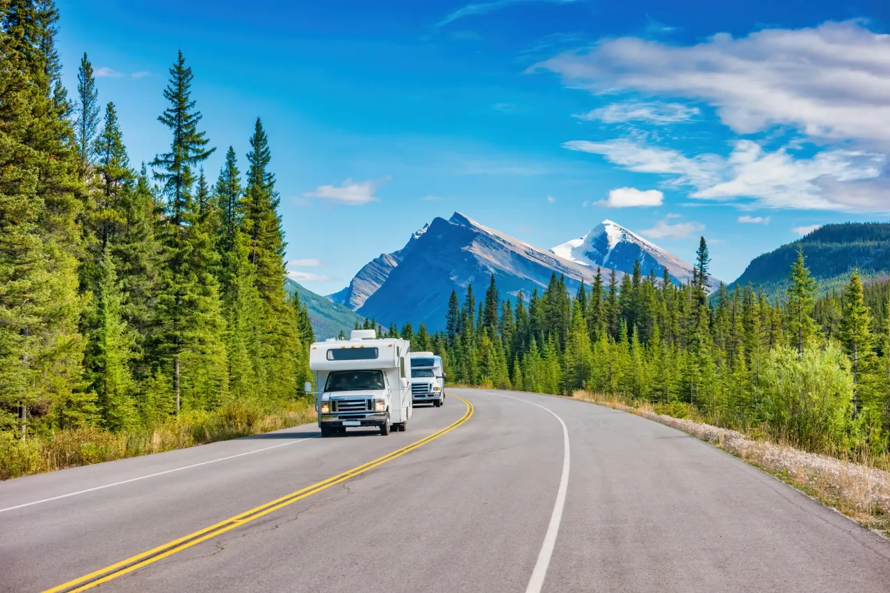 An RV on the road with trees on the side of the road and mountains off in the distance.