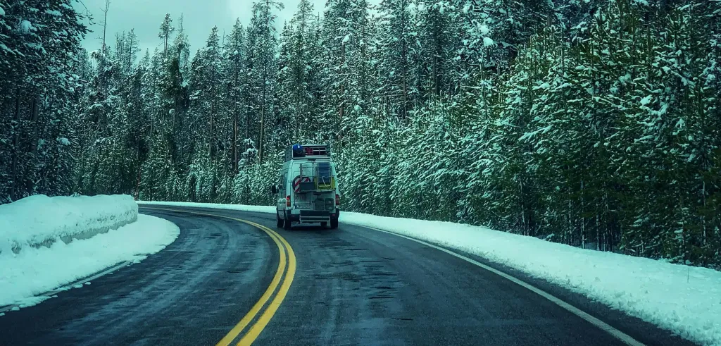 A sprinter van converted into a camper driving away on the road with snow on the ground and snow covered evergreen trees along the road.