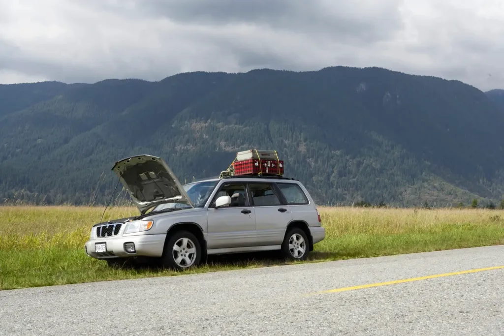 A silver SUV pulled over on the side of the road with the vehicle hood propped up. There is a dense green tree-covered mountain range in the distance.