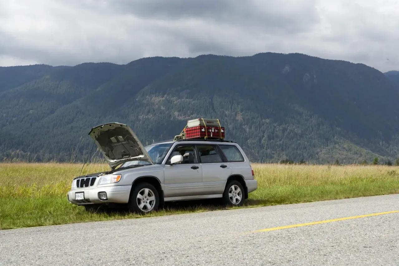 A silver SUV pulled over on the side of the road with the vehicle hood propped up. There is a dense green tree-covered mountain range in the distance.