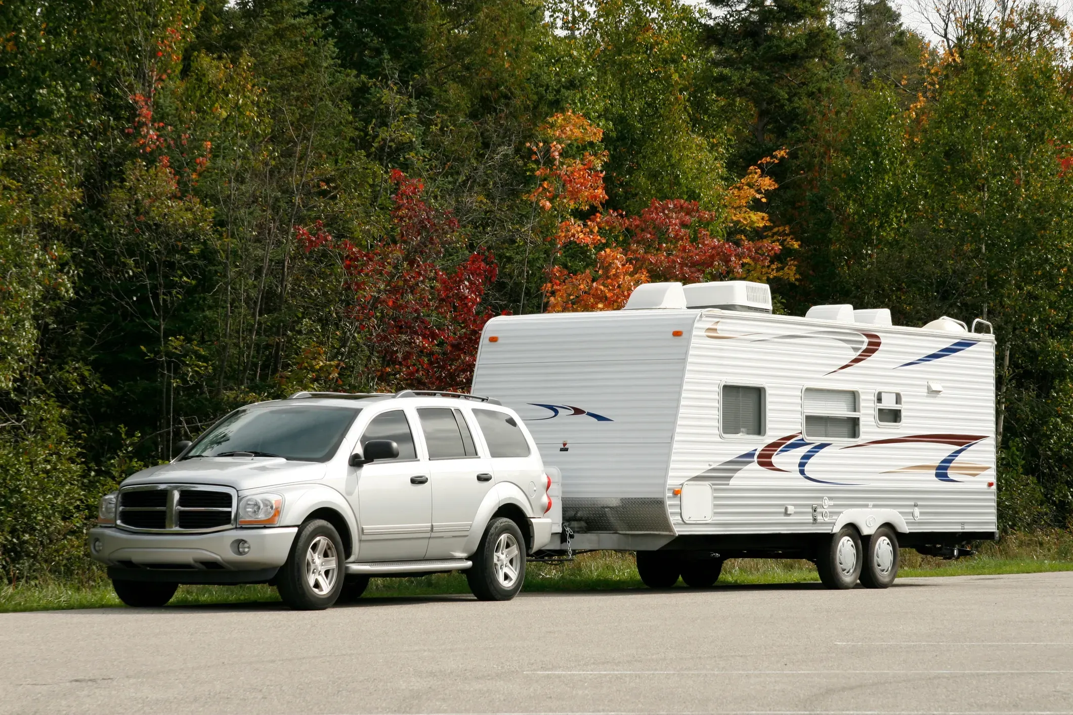 A silver SUV pulling a white travel trailer with trees in the background with some fall foliage.