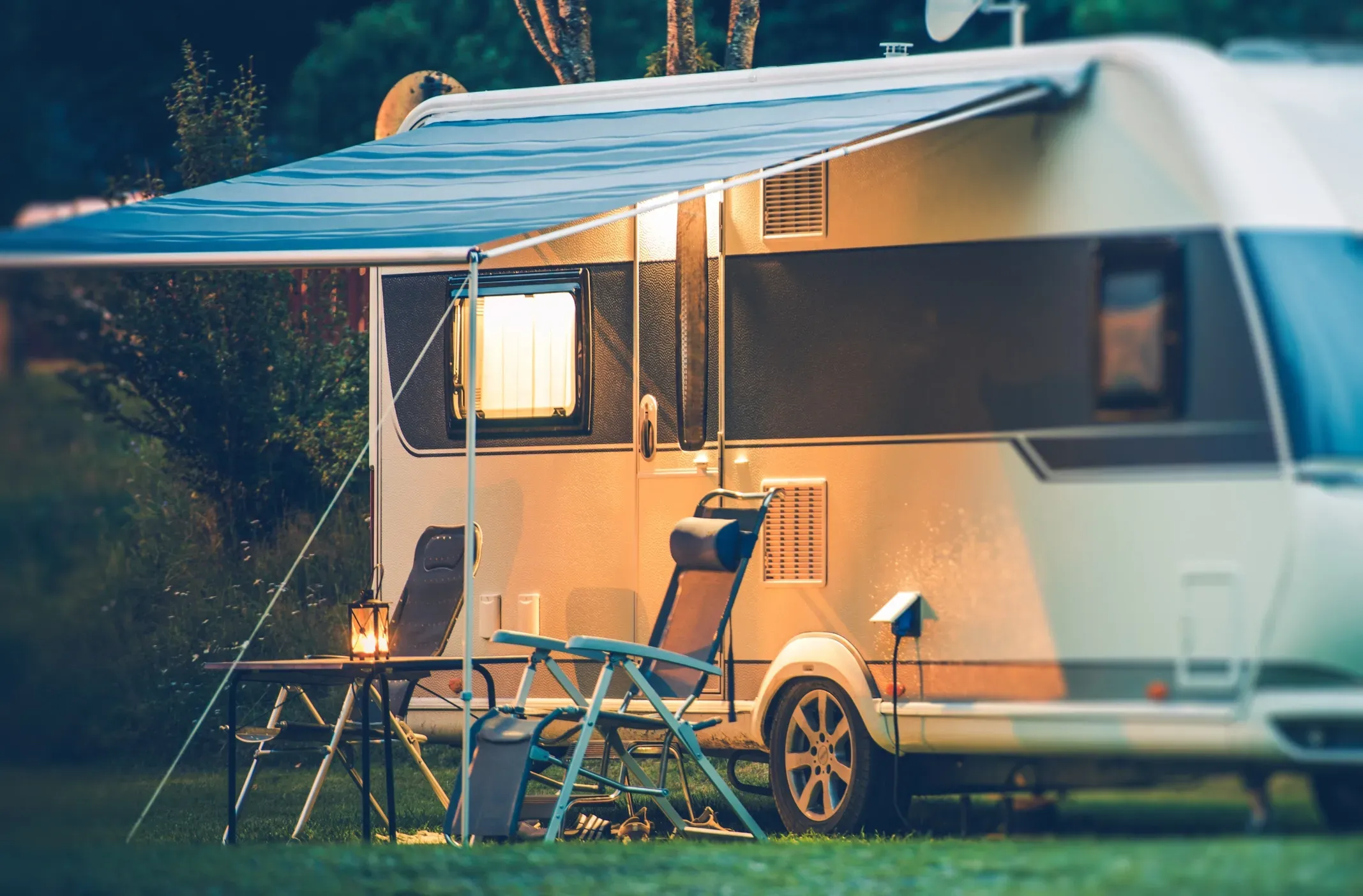 A travel trailer parked in a campsite with two camping chairs and a lantern on a table under the RV's awning.